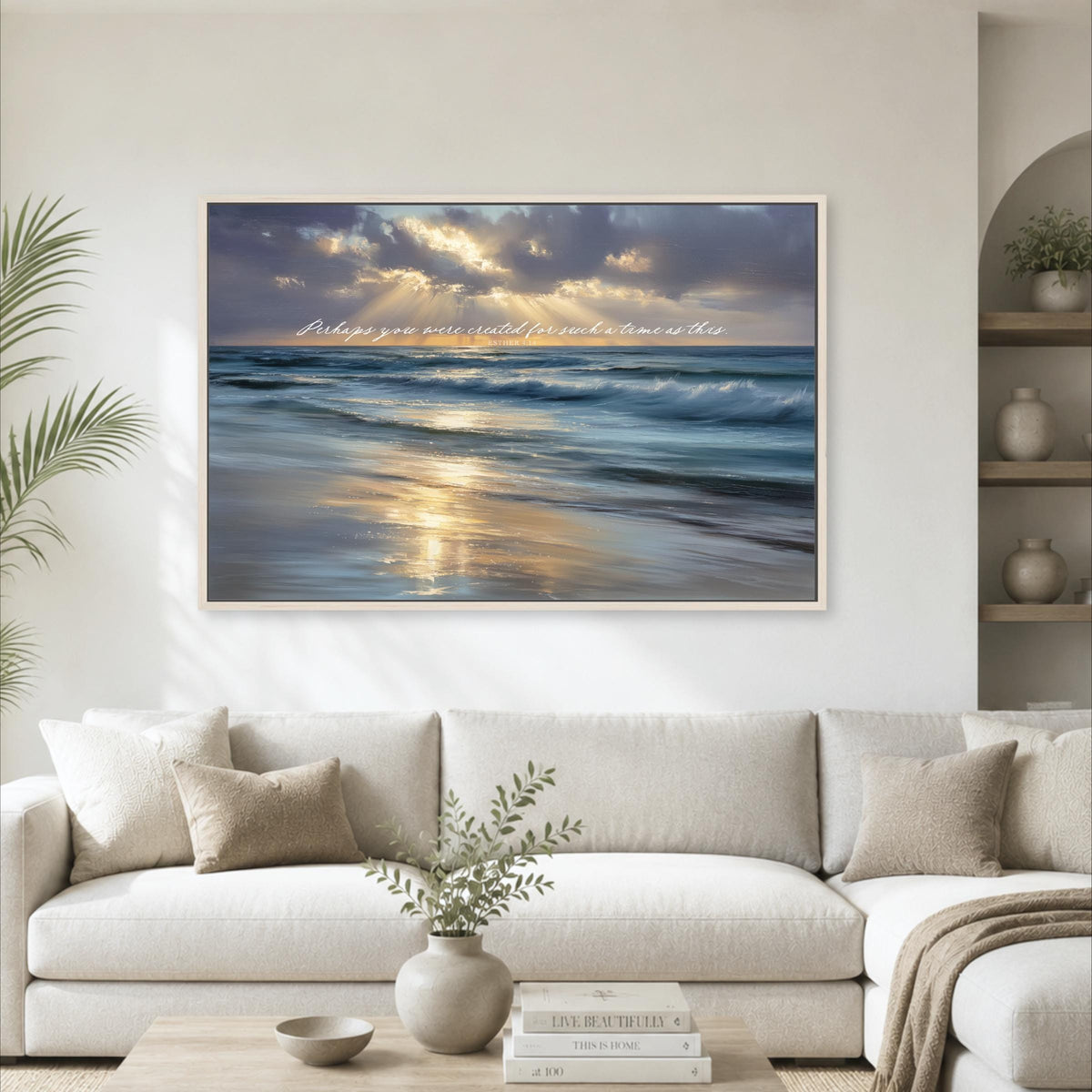 a large framed painting of a serene beach scene with waves crashing against the shore, set against a backdrop of a cloudy sky. The painting is hung on a wall above a white sofa, which is positioned in front of a coffee table. The room also includes a potted plant, a vase with greenery, and a bookshelf with decorative items.