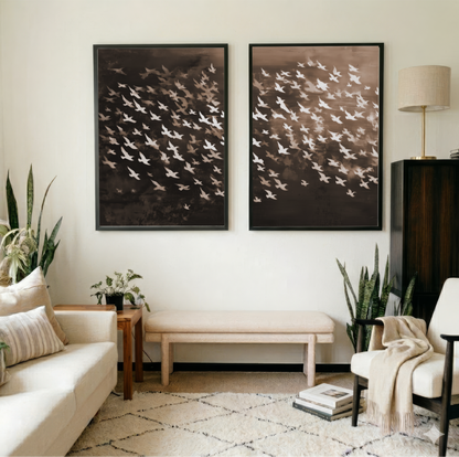 a living room with two large framed artworks hanging on the wall, depicting a flock of birds in flight. The room is furnished with a white sofa, a wooden side table, and a beige armchair. There are also several potted plants and a lamp in the room, contributing to the cozy and inviting atmosphere.