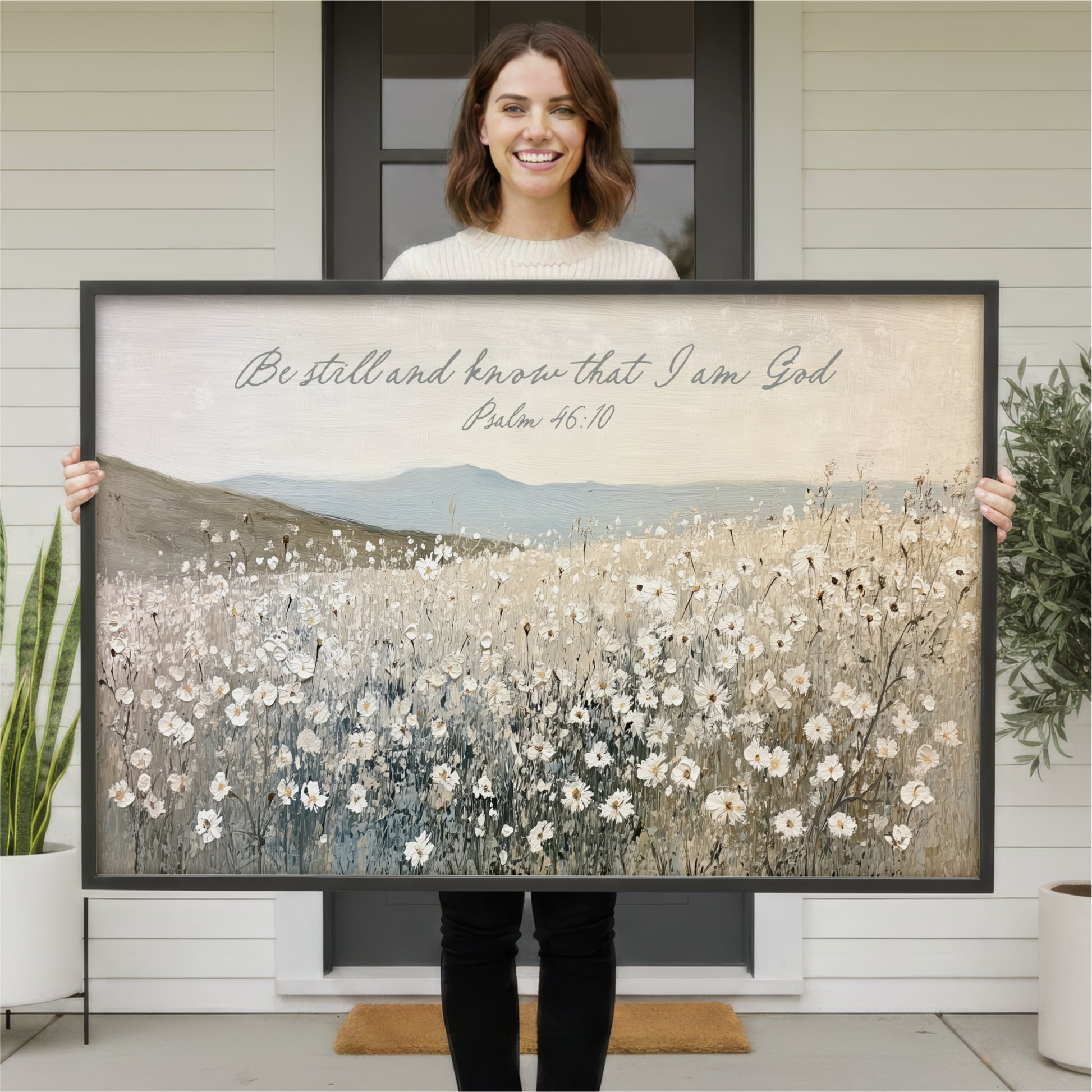 A woman is holding a large framed painting of a field of flowers in front of a house.