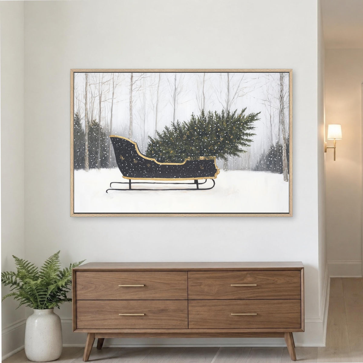 A framed painting of a black sled carrying a large Christmas tree is displayed on a white wall above a wooden dresser.