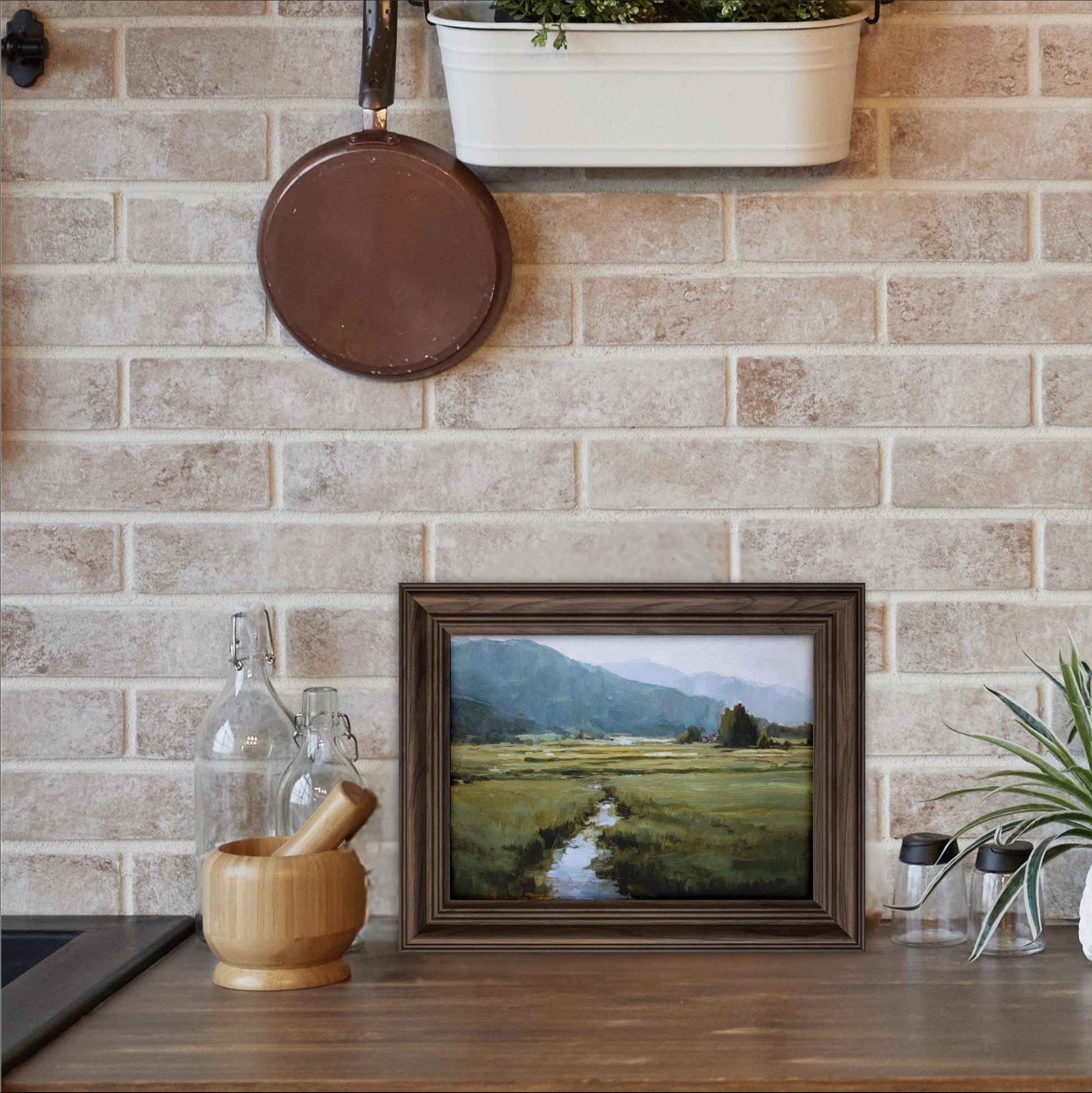 a framed landscape painting hanging on a brick wall above a wooden table with various kitchen items, including a wooden bowl, a mortar and pestle, and a plant.