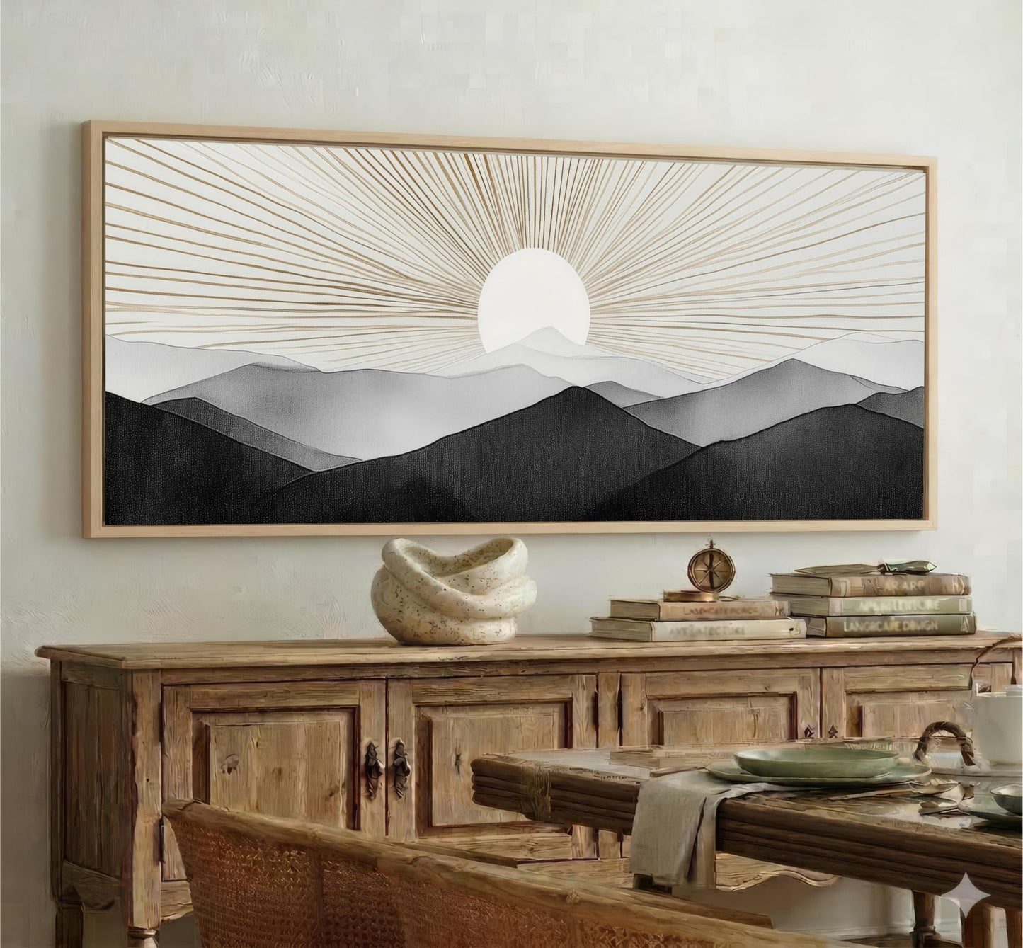 a large framed artwork depicting a serene landscape with mountains and a sun-like design in the sky. The artwork is mounted on a wall above a wooden cabinet, which has a vase and some books on top. The overall setting appears to be a cozy and inviting living space.