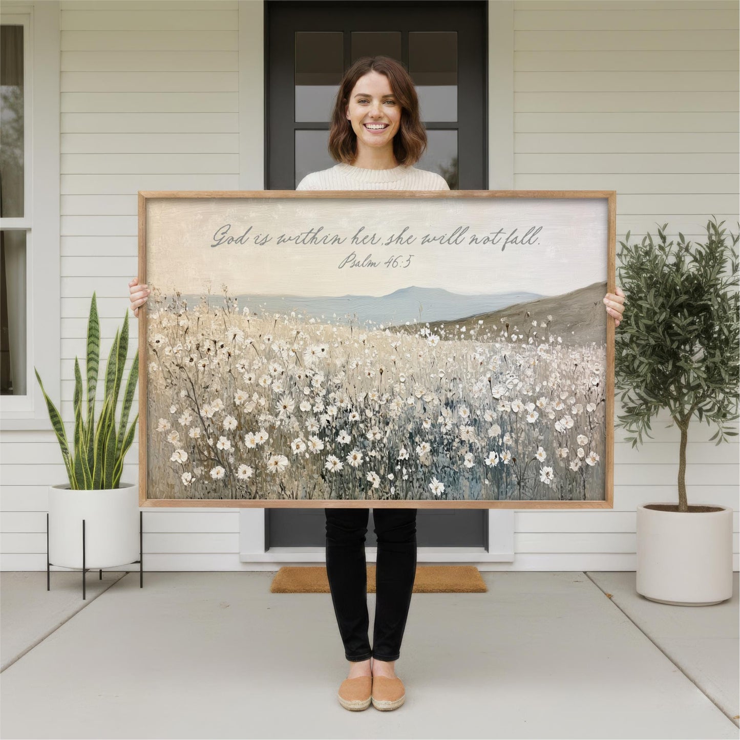A woman stands in front of a white house, holding a large framed print of a field of flowers.