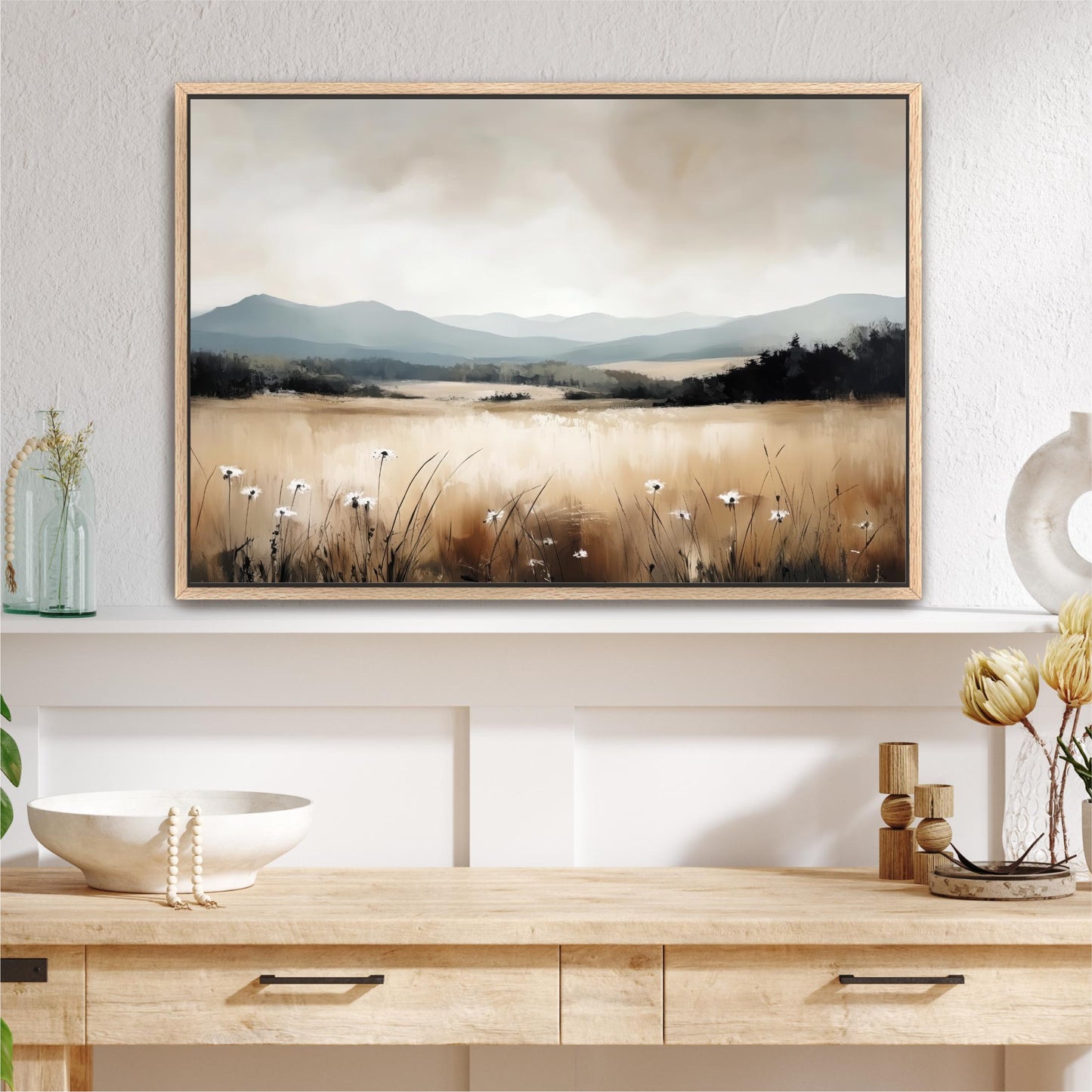 a large framed painting of a field with flowers hanging on a wall above a wooden table with a bowl and a vase.