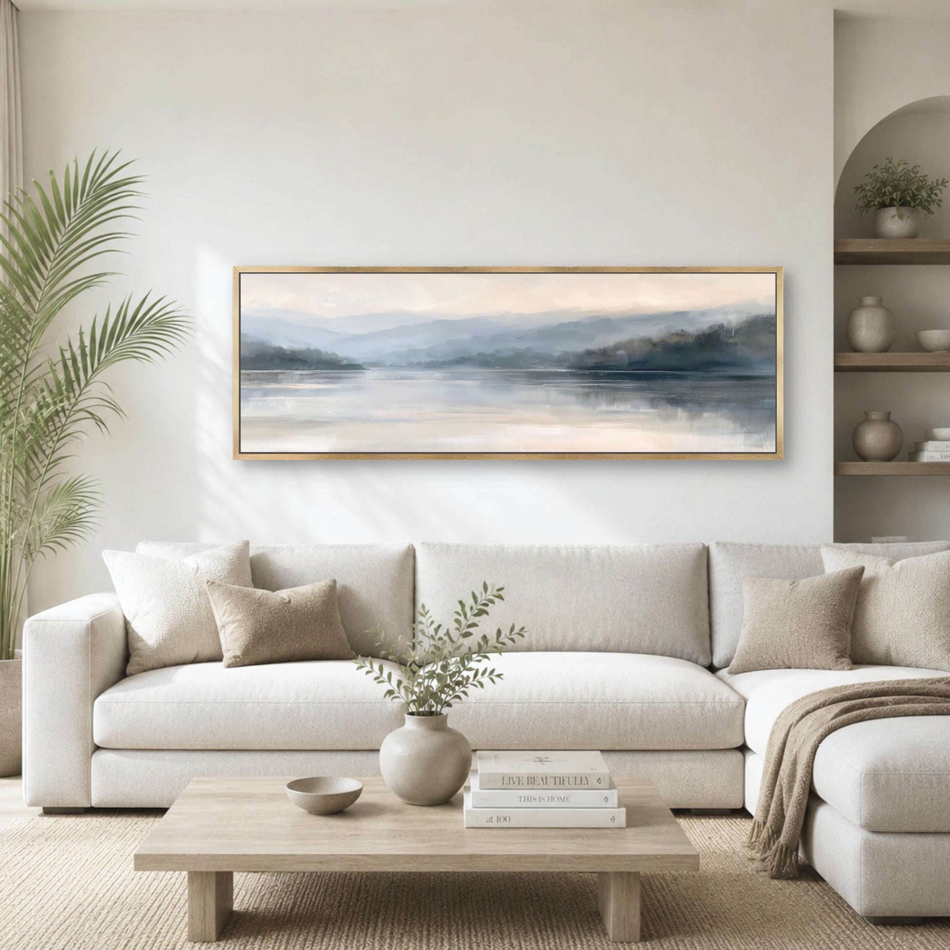 a serene living room with a large, framed landscape painting hanging on the wall above a white sofa. The painting features a tranquil scene of a lake surrounded by mountains, creating a peaceful and calming atmosphere. The room is decorated with a potted plant, a vase with greenery, and a wooden coffee table with books and a bowl.