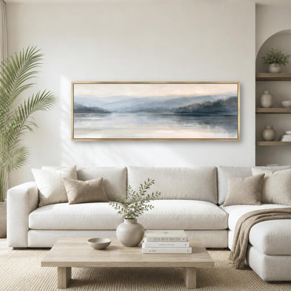 a serene living room with a large, framed landscape painting hanging on the wall above a white sofa. The painting features a tranquil scene of a lake surrounded by mountains, creating a peaceful and calming atmosphere. The room is decorated with a potted plant, a vase with greenery, and a wooden coffee table with books and a bowl.