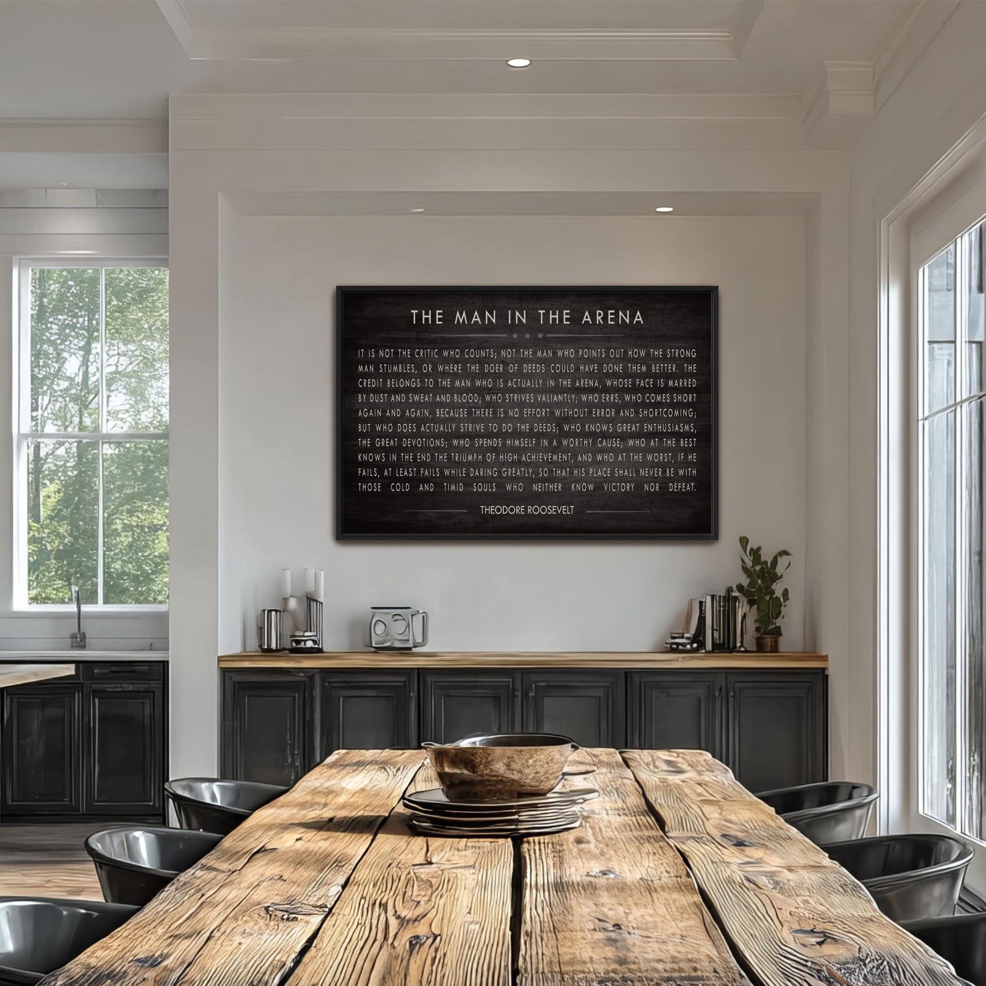 a large black framed text on a white wall, which reads "THE MAN IN THE ARENA" and is accompanied by a quote. The text is in a cursive font and is centrally positioned above a wooden dining table with chairs. The dining table is situated in a room with a window and a bookshelf, creating a cozy and inviting atmosphere.