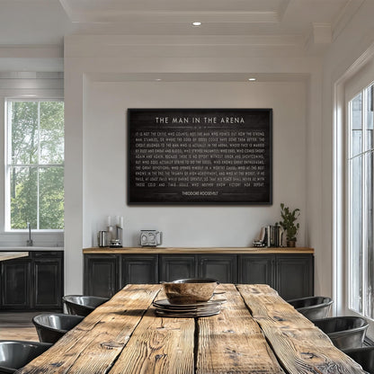 a large black framed text on a white wall, which reads "THE MAN IN THE ARENA" and is accompanied by a quote. The text is in a cursive font and is centrally positioned above a wooden dining table with chairs. The dining table is situated in a room with a window and a bookshelf, creating a cozy and inviting atmosphere.