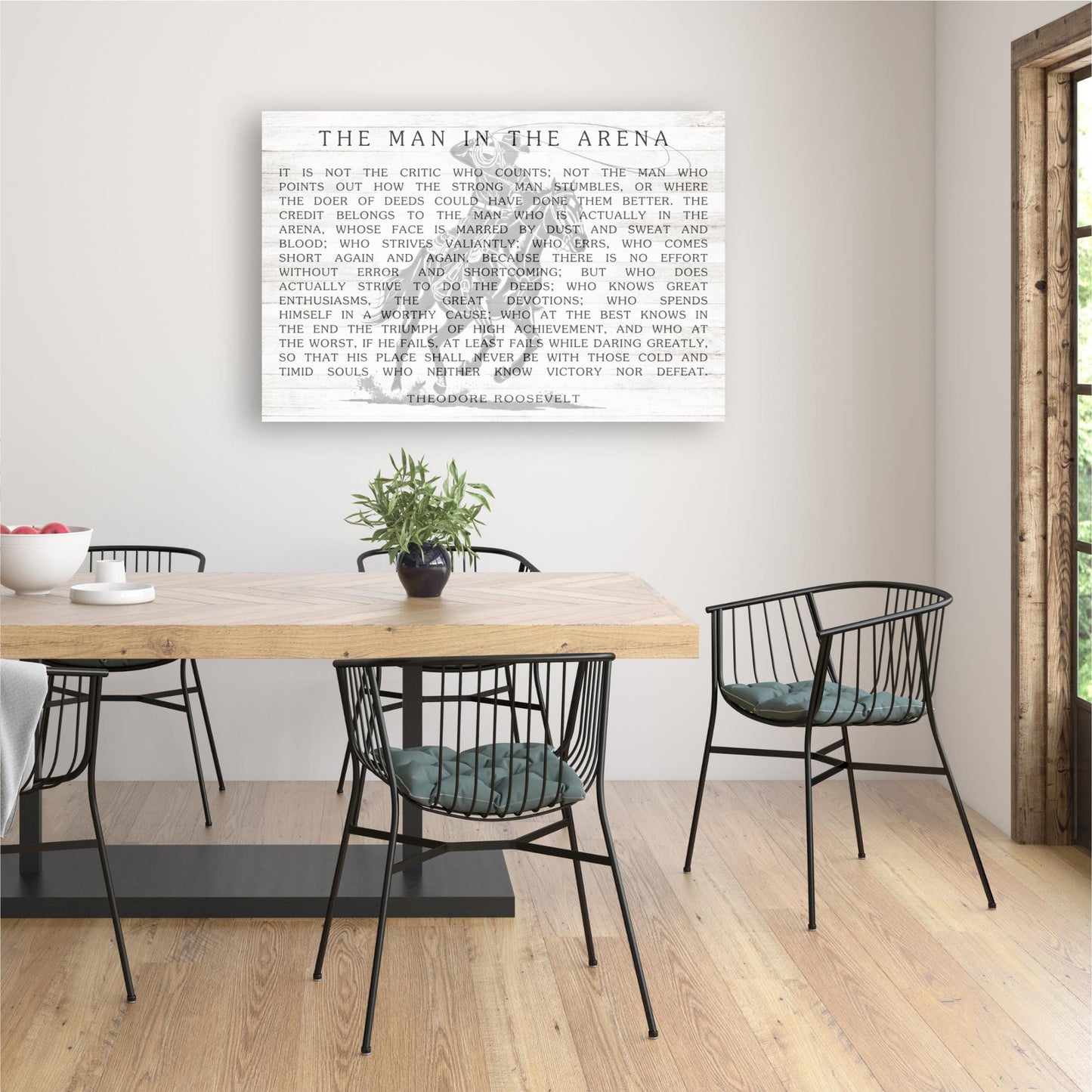 a dining room with a wooden table and black metal chairs. Above the table, there is a framed quote in white text that reads "The man in the arena".