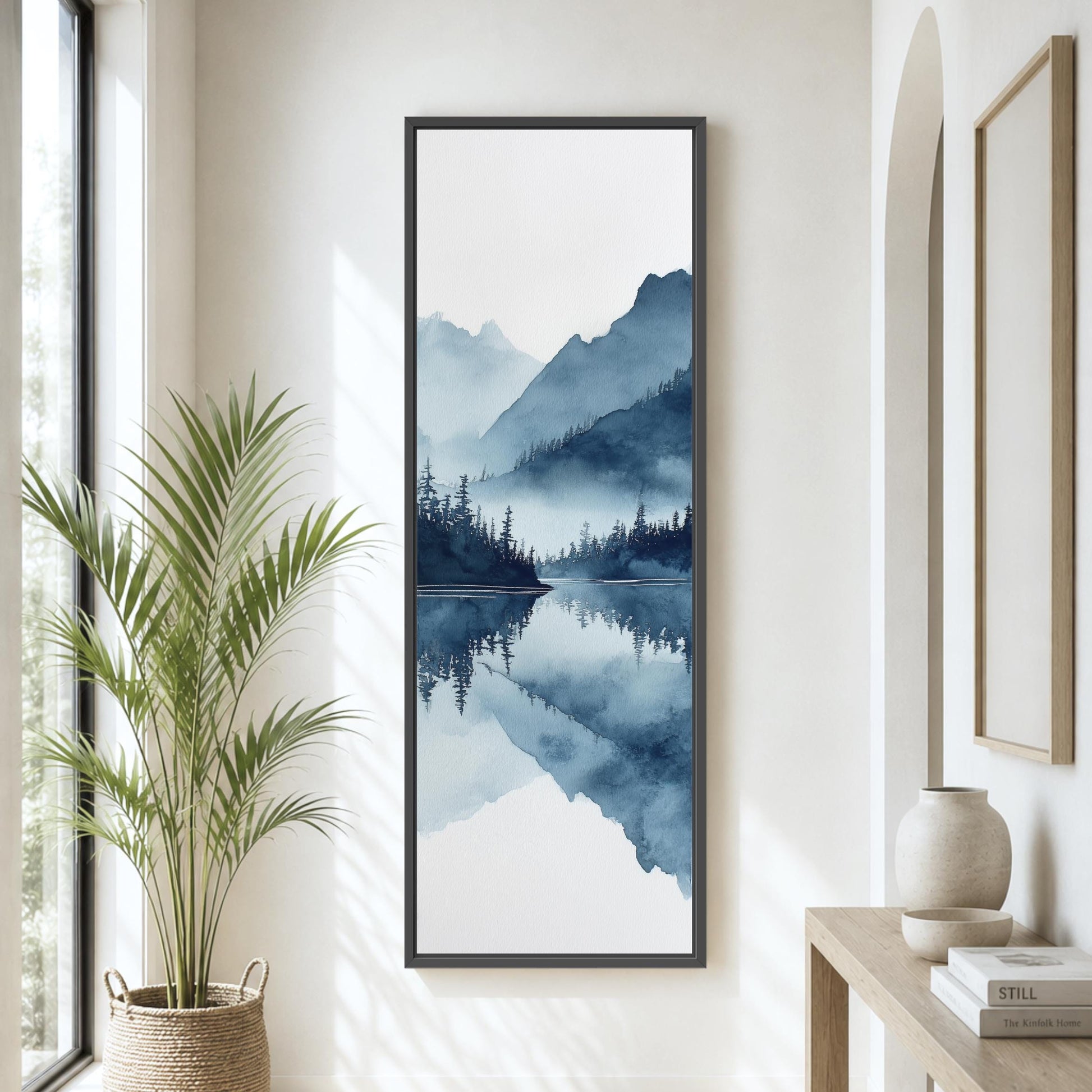 a large framed painting of a serene landscape with mountains, trees, and a lake, hanging on a wall in a room with a potted plant and a wooden table.