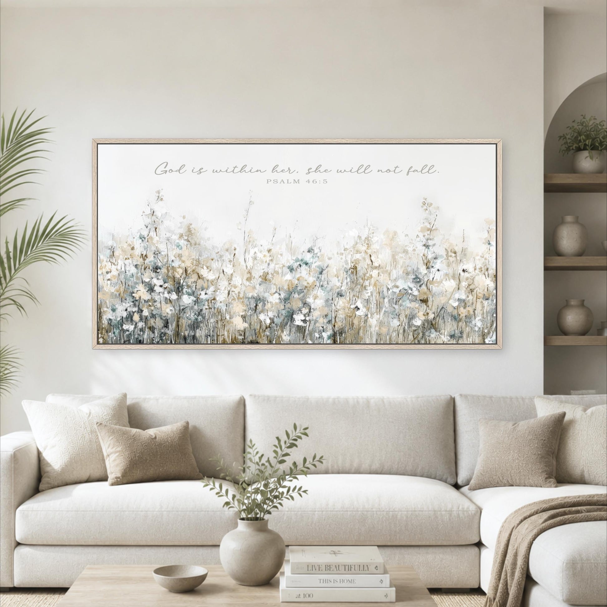 A living room with a large framed print of a floral landscape hanging on the wall above a white sofa. The print features a variety of flowers in different shades of white, blue, and green. The sofa is adorned with throw pillows and a vase of greenery. The room also includes a coffee table with books and decorative items, creating a cozy and inviting atmosphere.