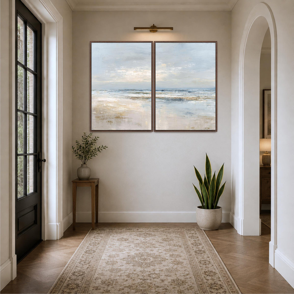 a hallway with a large painting of a beach scene hanging on the wall. The painting is framed in brown and is placed above a small table with a plant on it. The hallway has a wooden floor and a rug in front of the table. There is also a doorway on the right side of the hallway.
