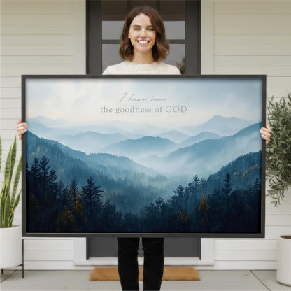 A woman is holding a large framed print of a mountain landscape with the text "I have seen the goodness of GOD" written on it.