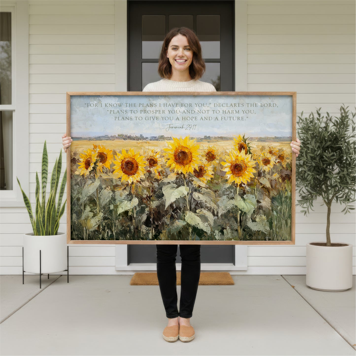 A woman stands in front of a white house, holding a large painting of a field of sunflowers.