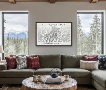 a cozy living room with a large window overlooking a scenic mountain landscape. The room is furnished with a gray sectional sofa adorned with red and white plaid throw pillows, and a wooden coffee table with a teapot and two mugs. Above the sofa, a framed quote is displayed, and the room is decorated with various books and other items, creating a warm and inviting atmosphere.
