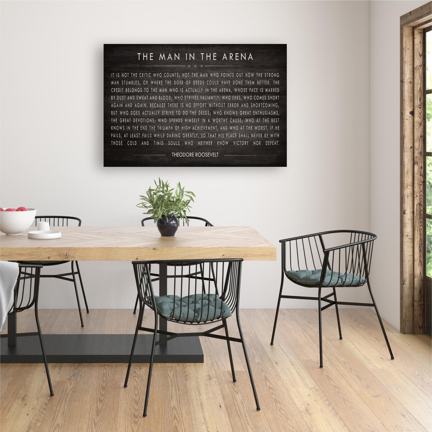 a dining room with a wooden table and black metal chairs. Above the table, there is a black and white framed quote that reads "The man in the arena".