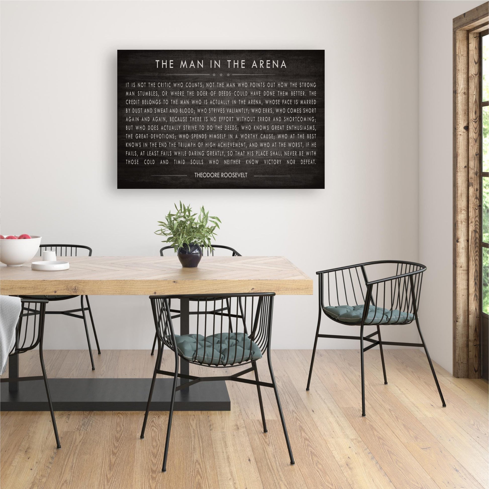 a dining room with a wooden table and black metal chairs. Above the table, there is a black and white framed quote that reads "The man in the arena".