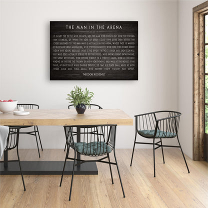 a dining room with a wooden table and black metal chairs. Above the table, there is a black and white framed quote that reads "The man in the arena".