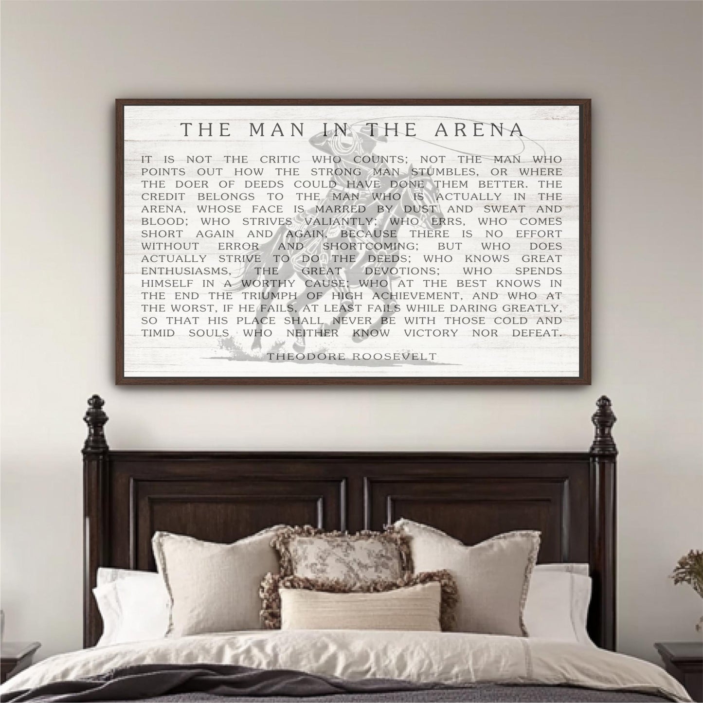 a framed quote by Theodore Roosevelt, "The Man in the Arena", displayed on a wall above a bed with white linens and a wooden headboard.