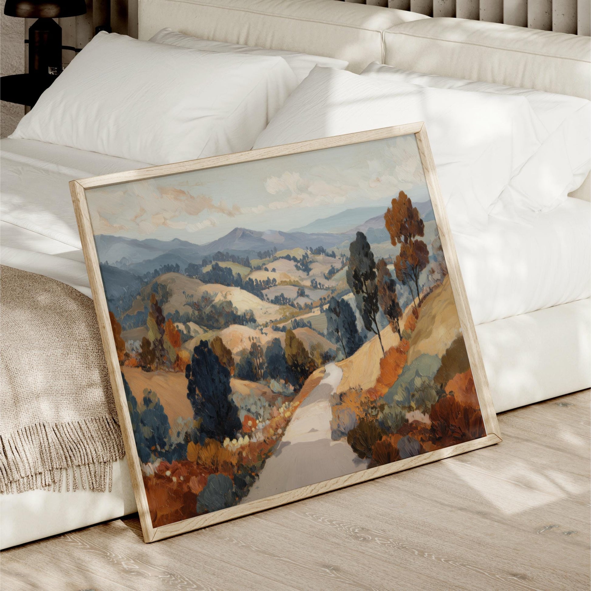 a large landscape painting with a winding road and trees in the foreground, displayed on a wooden floor next to a white bed.