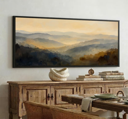 a large framed landscape painting hanging on a wall above a wooden cabinet. The painting depicts a serene mountain scene with a warm, golden glow. The cabinet below the painting has a vase and some books on top, creating a cozy and inviting atmosphere.