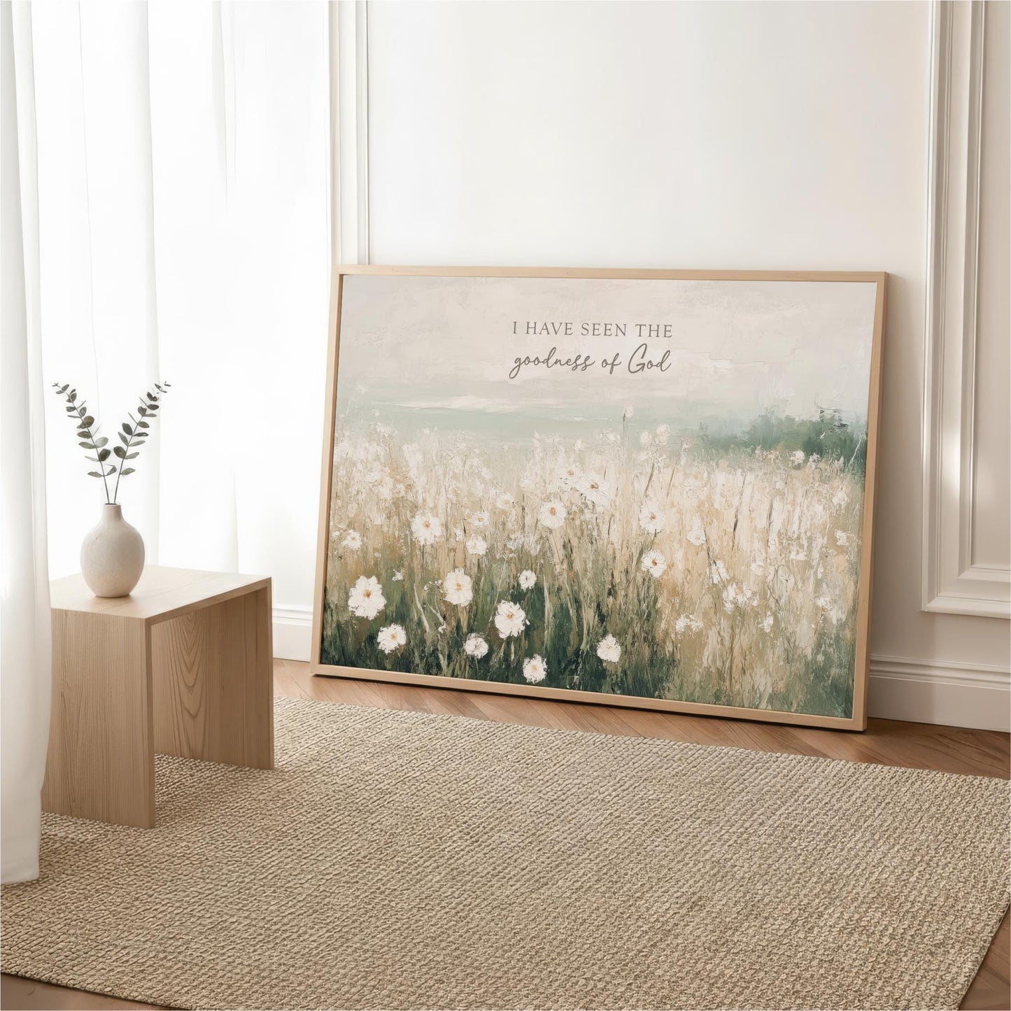 a large framed painting of a field of white flowers, with a vase of greenery on a small wooden table in the foreground.