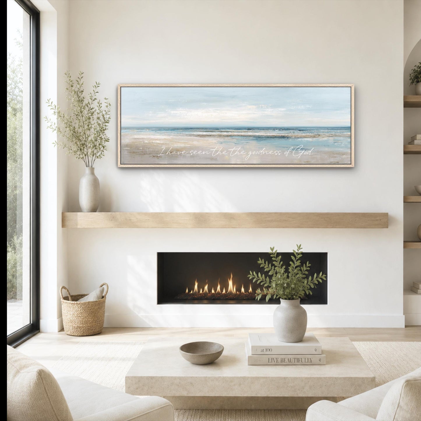 a cozy living room with a fireplace, a coffee table, and a framed landscape painting above it. The painting features a serene beach scene with a clear blue sky and calm ocean. The room is decorated with a vase of greenery, a woven basket, and a stack of books.