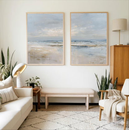 a cozy living room with two large paintings of a serene beach scene hanging on the wall. The room is furnished with a white sofa, a wooden side table, and a wooden cabinet. The space is decorated with potted plants and a rug, creating a warm and inviting atmosphere.