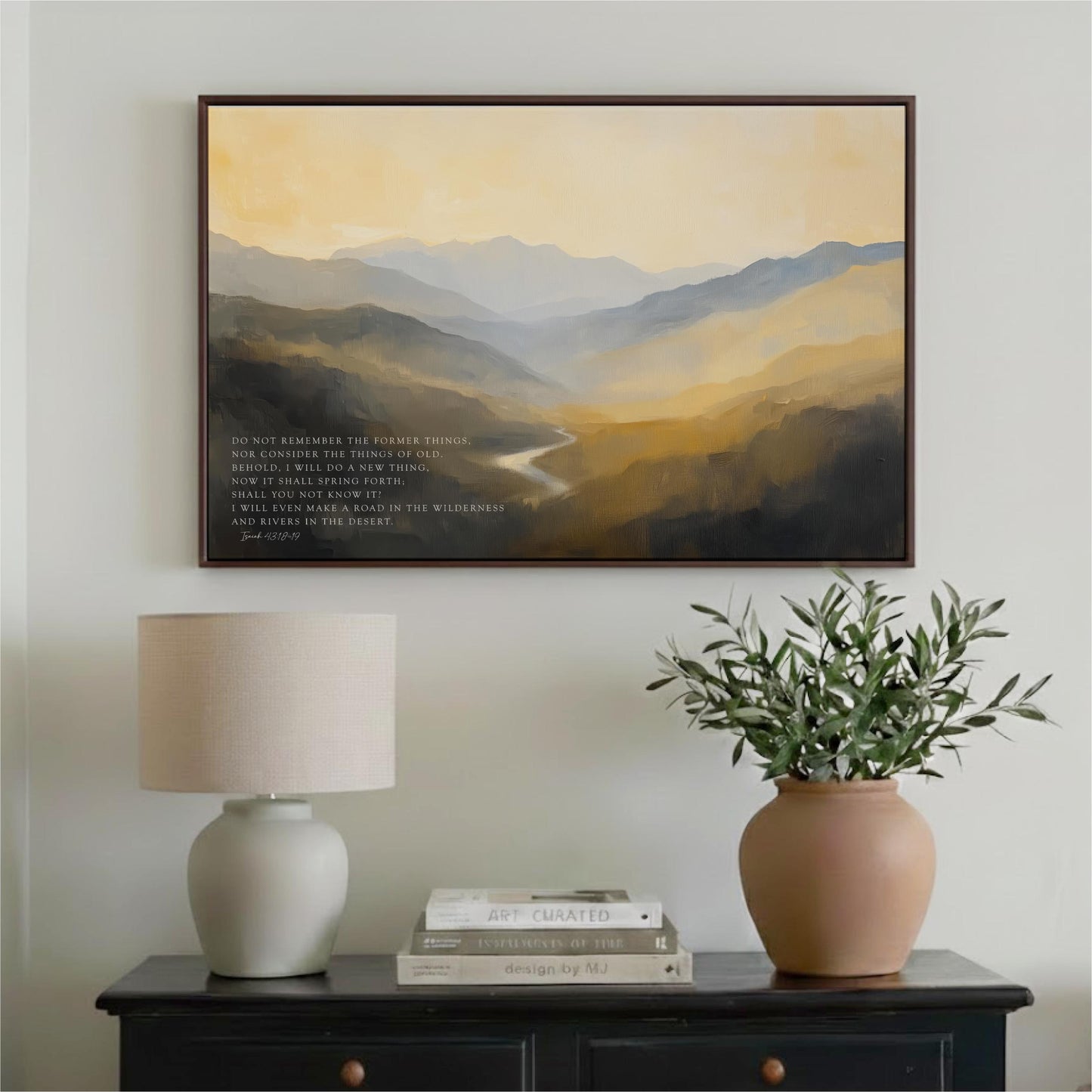 a framed landscape painting hanging on a wall above a black table with a lamp and a vase of greenery.