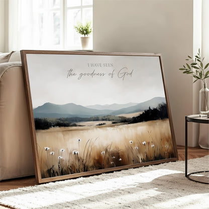 a large framed print of a serene landscape with mountains, grass, and wildflowers, placed on a wooden floor in a living room.