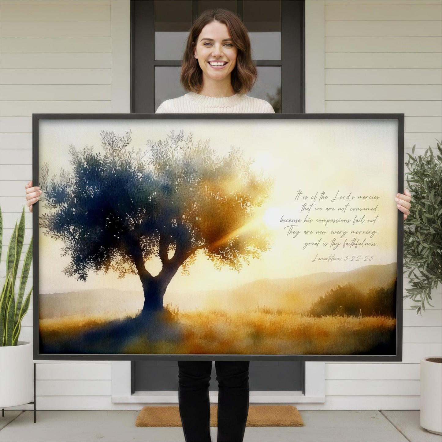A woman is holding a large framed print of a serene landscape with a tree and a sunset in the background.