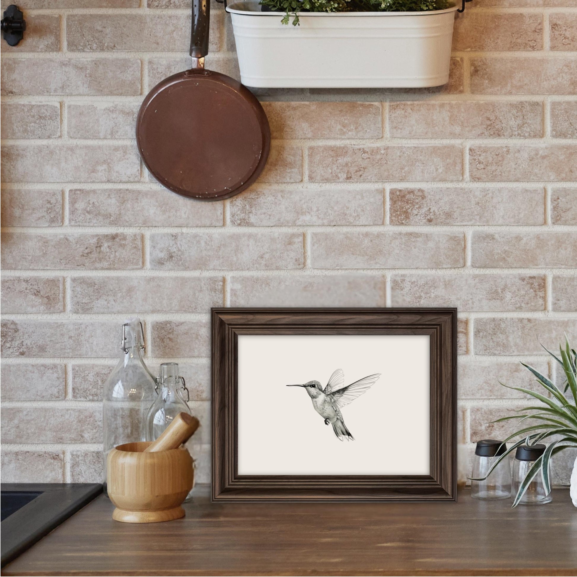 a framed print of a hummingbird on a wooden surface, with a pot plant and various kitchen items such as a wooden bowl, a glass bottle, and a wooden mortar and pestle nearby.