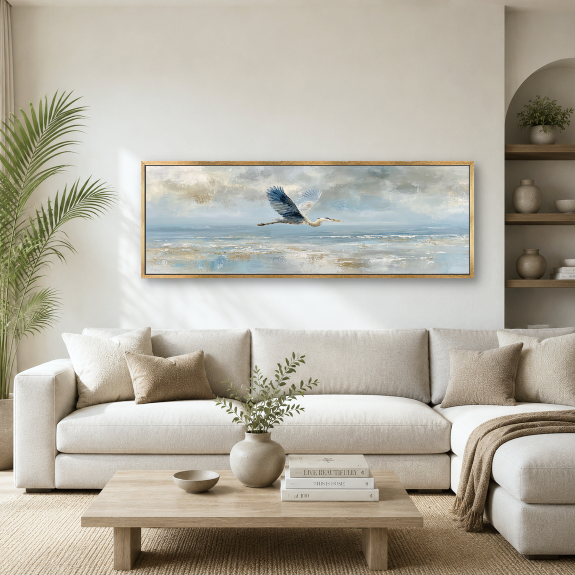 a serene living room with a large painting of a bird in flight hanging on the wall above a white sofa. The painting is framed in gold and features a blue bird soaring over a tranquil body of water. The room is decorated with a potted plant, a vase with greenery, and a wooden coffee table with books and a bowl.
