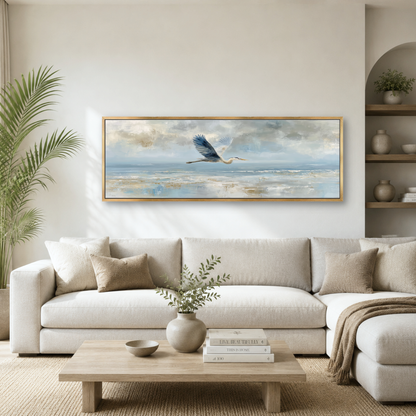 a serene living room with a large painting of a bird in flight hanging on the wall above a white sofa. The painting is framed in gold and features a blue bird soaring over a tranquil body of water. The room is decorated with a potted plant, a vase with greenery, and a wooden coffee table with books and a bowl.