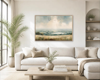 a cozy living room with a large, framed landscape painting hanging on the wall above a white sofa. The painting features a serene countryside scene with rolling hills and a tranquil body of water. The room is decorated with various plants, vases, and books, creating a warm and inviting atmosphere.