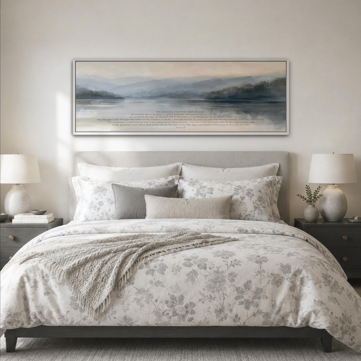a serene bedroom scene with a large bed adorned with a floral comforter and pillows, flanked by two nightstands with lamps. Above the bed, a large framed landscape painting hangs on the wall, depicting a tranquil body of water surrounded by mountains.