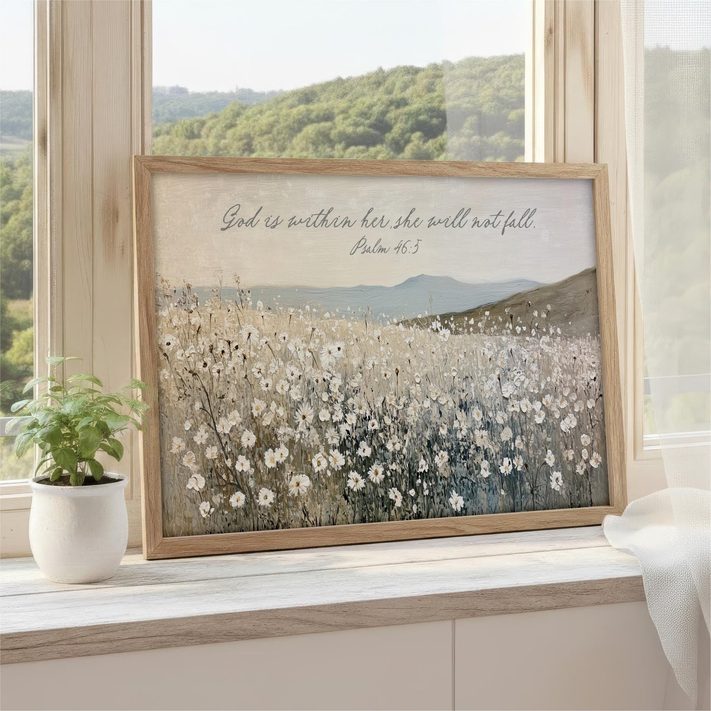 A framed painting of a field of flowers is displayed on a windowsill, with a potted plant to the left and a scenic view of a mountain range visible through the window.