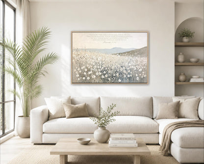 a serene living room with a large painting of a field of flowers hanging on the wall above a white sofa. The sofa is adorned with throw pillows and a vase of greenery. A coffee table sits in front of the sofa, and a bookshelf filled with books and decorative items can be seen in the background.