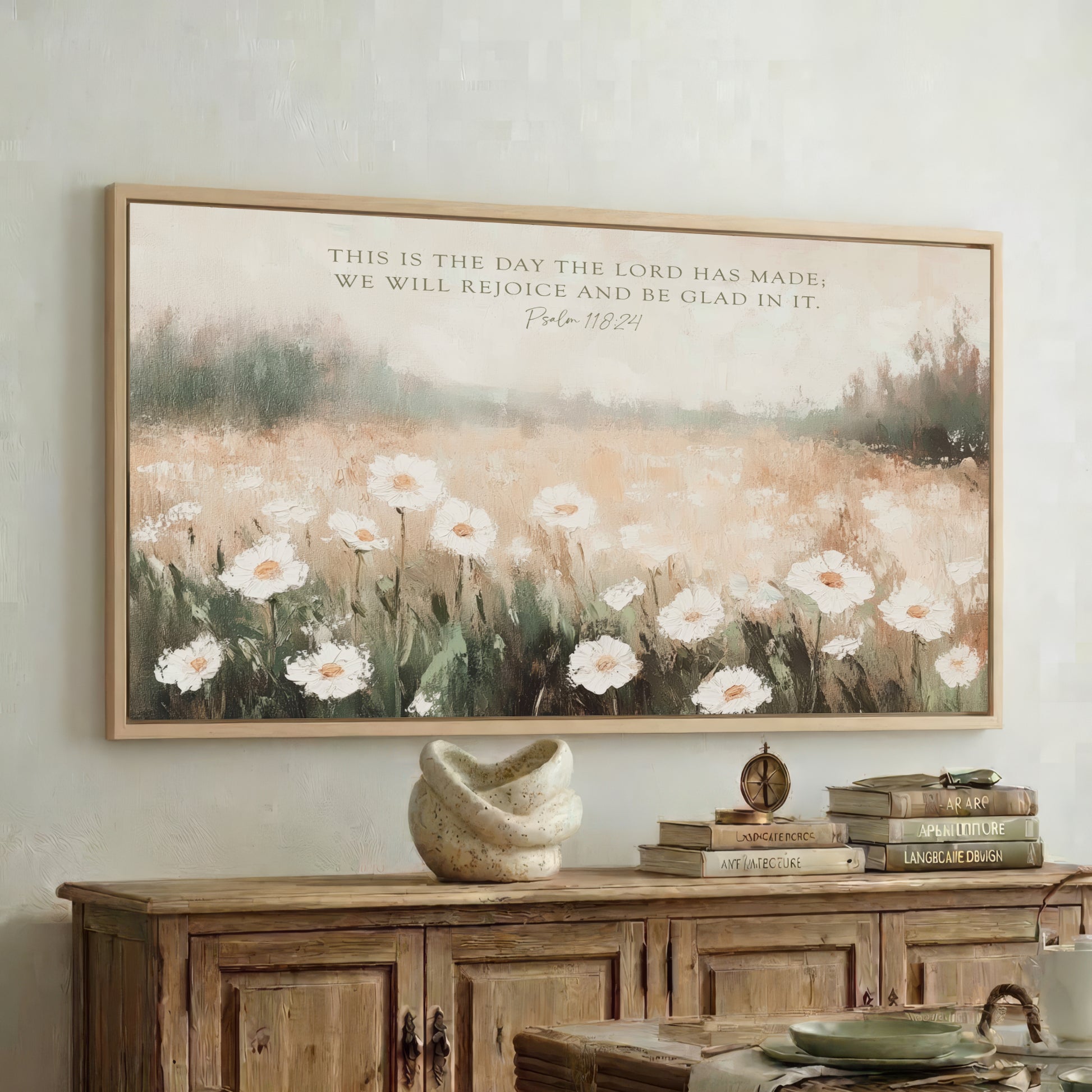 a framed painting of a field of white flowers hanging on a wall above a wooden cabinet. The cabinet has a stack of books on top of it.