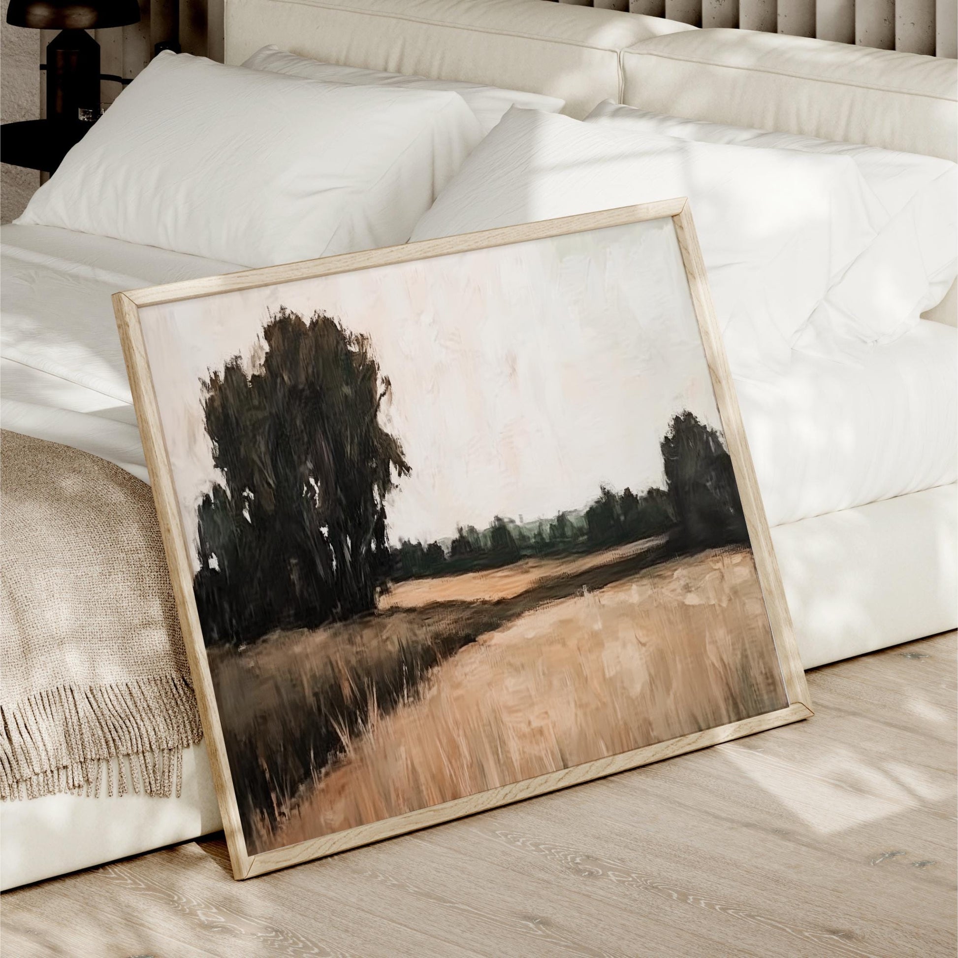 a painting of a field with trees in the background, displayed on a wooden floor next to a white bed.