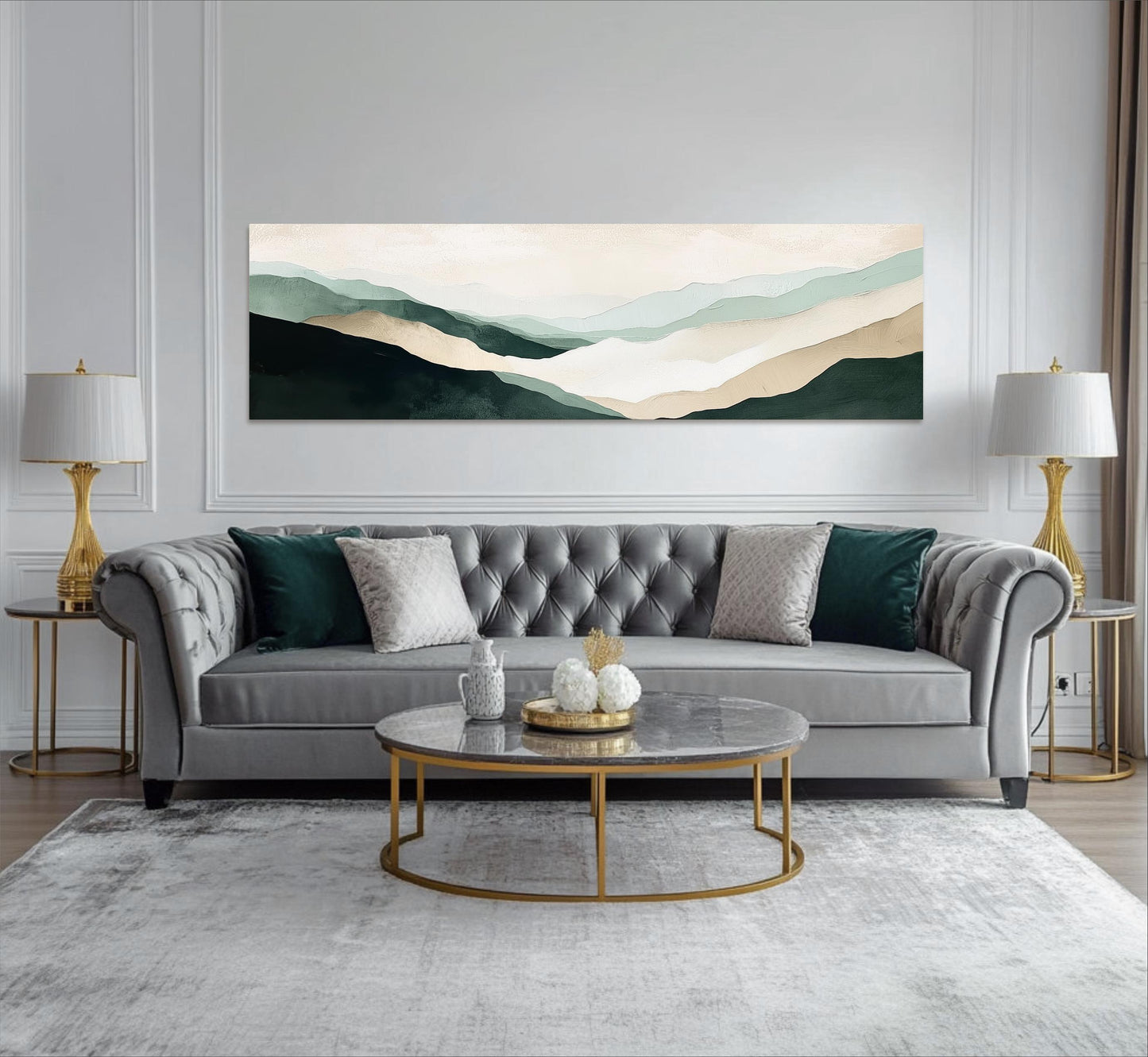 a modern living room with a gray tufted sofa, a round marble coffee table, and two gold side tables with lamps. Above the sofa, there is a large landscape painting depicting a mountain range with a light blue sky.
