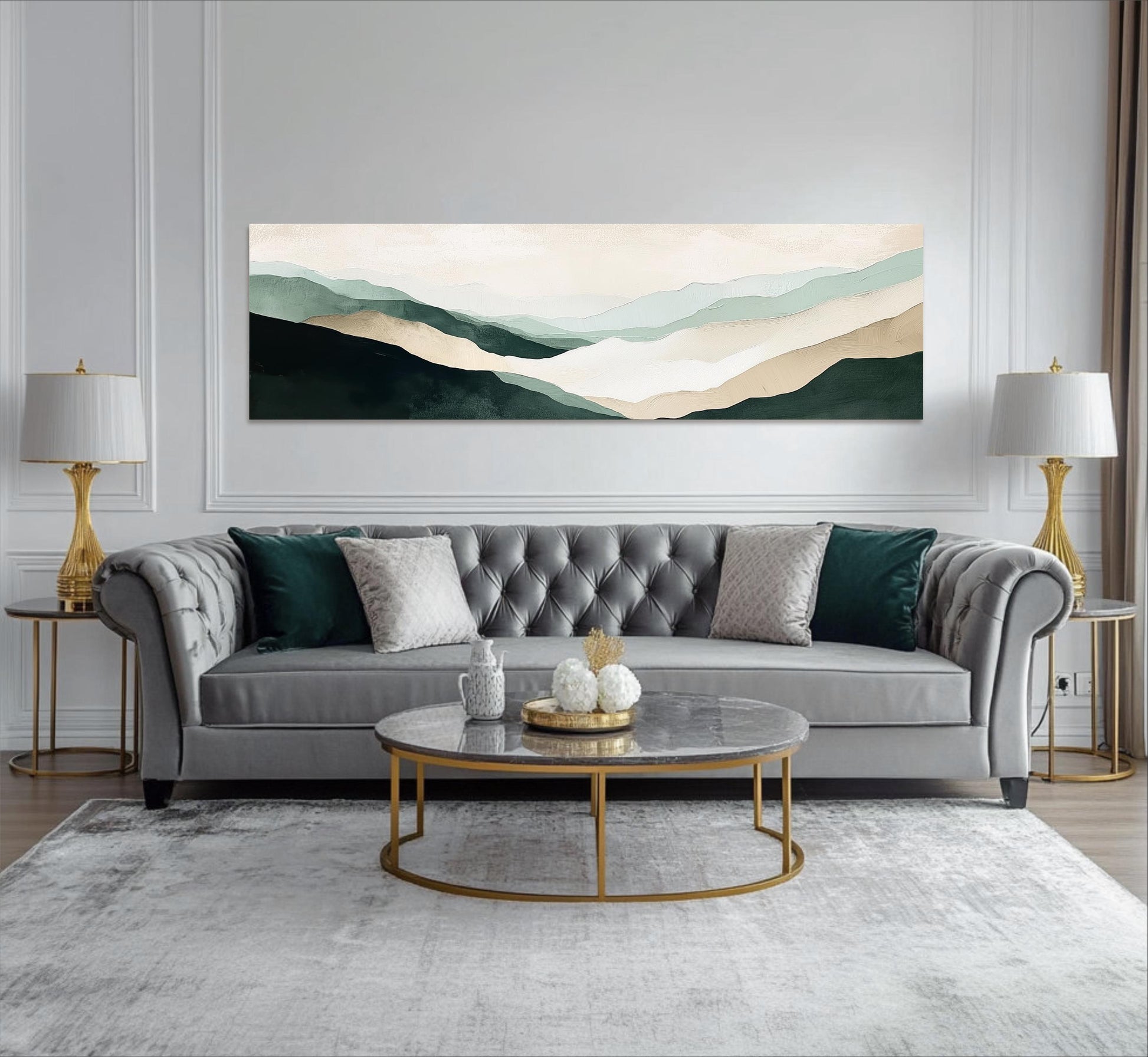 a modern living room with a gray tufted sofa, a round marble coffee table, and two gold side tables with lamps. Above the sofa, there is a large landscape painting depicting a mountain range with a light blue sky.