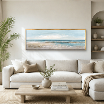 a living room with a large framed painting of a beach scene hanging on the wall above a white sofa. The painting depicts a serene beach landscape with a clear blue sky and calm ocean. The room is decorated with various items, including a potted plant, a vase with greenery, and a stack of books. The overall atmosphere of the room is calm and inviting, with the beach scene as the focal point.