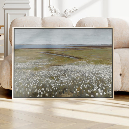 a large framed painting of a field with white flowers, displayed on a wooden floor in front of a couch.