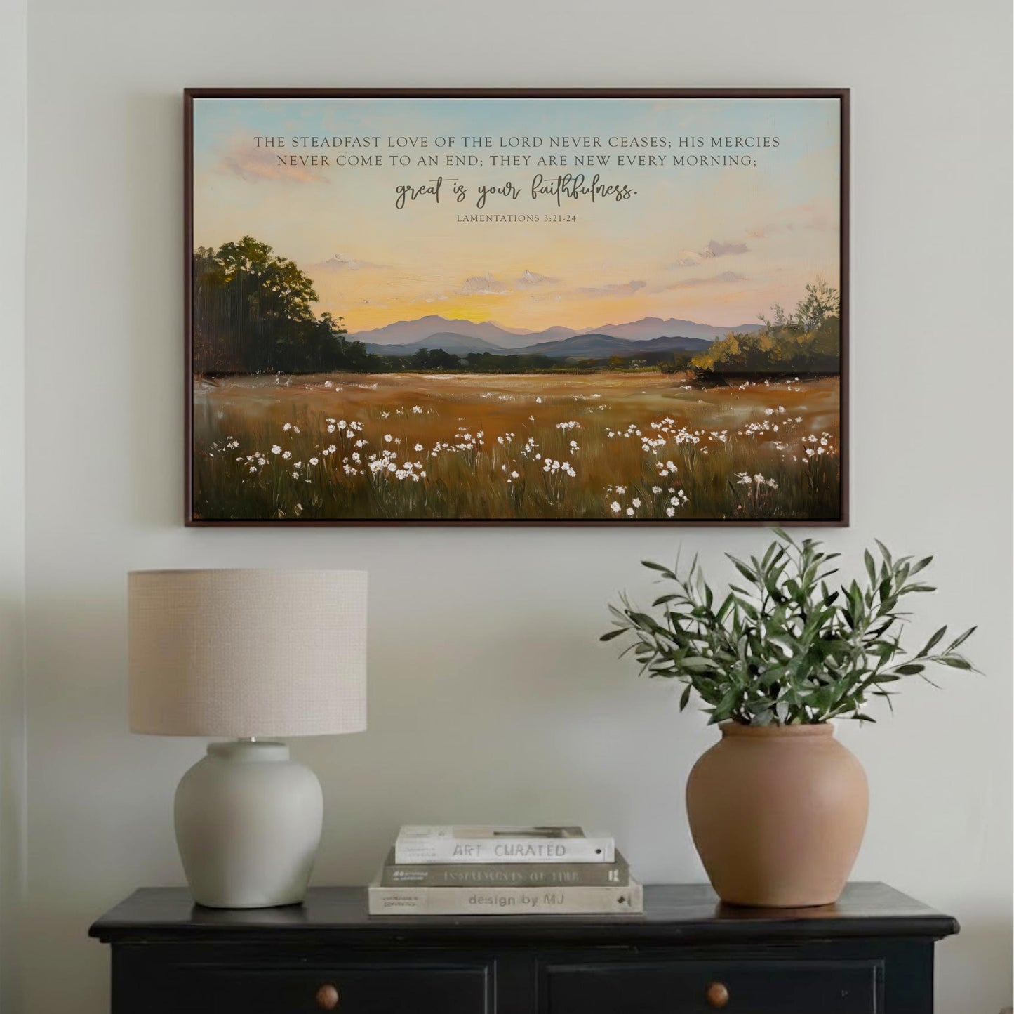 a framed painting of a serene landscape with a field of white flowers and a sunset sky, hanging on a wall above a wooden table with a lamp and a potted plant.