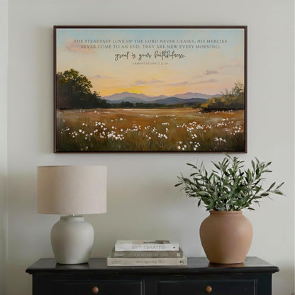 a framed painting of a serene landscape with a field of white flowers and a sunset sky, hanging on a wall above a wooden table with a lamp and a potted plant.