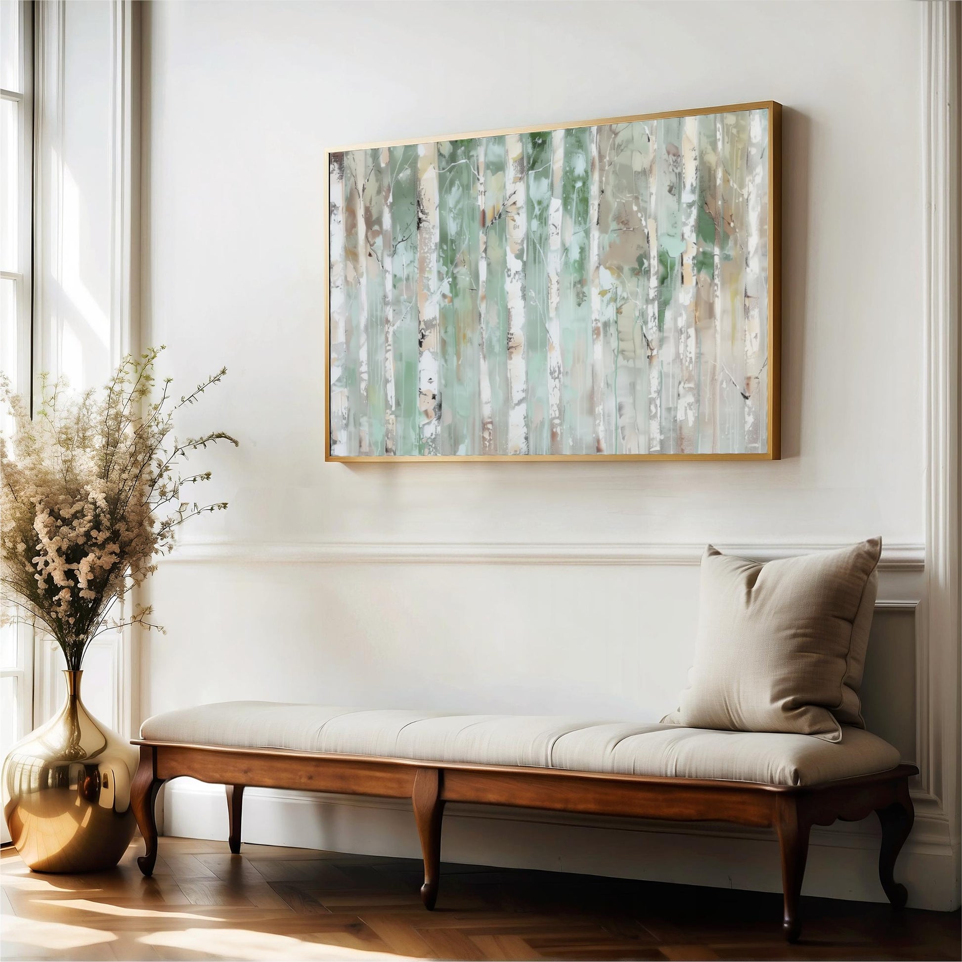 a large painting of trees hanging on a white wall above a wooden bench with a beige cushion. The painting is framed in gold and has a green and white color scheme. The bench is placed in front of a window, and there is a vase of flowers on the floor next to it.