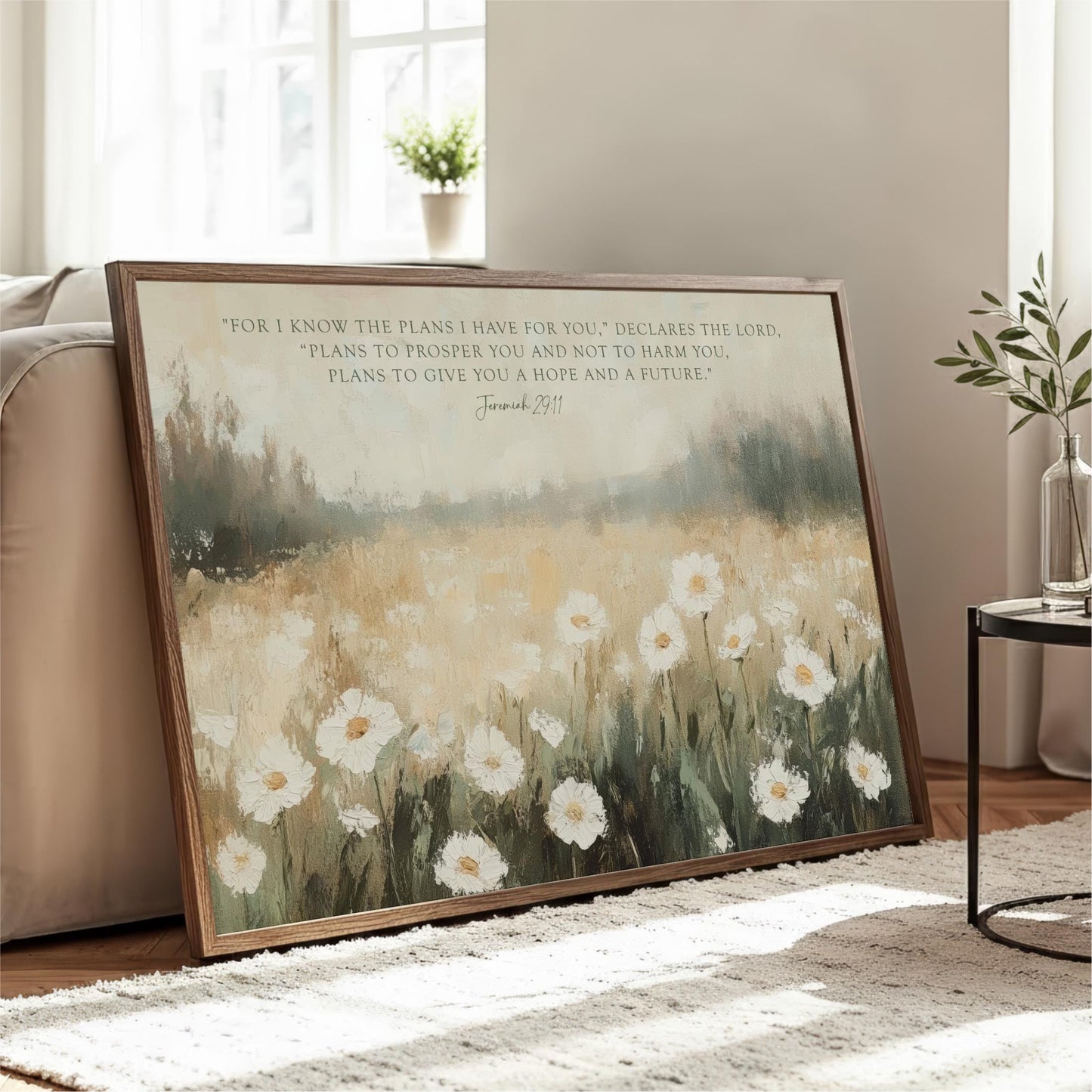 A framed painting of a field of white flowers is displayed on a wooden floor in a living room, with a potted plant and a vase nearby.