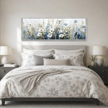 a serene bedroom scene with a large bed adorned with a floral comforter and pillows, flanked by two nightstands with lamps. Above the bed, a large painting of a field of flowers in various shades of blue and white adds a touch of nature and tranquility to the space.