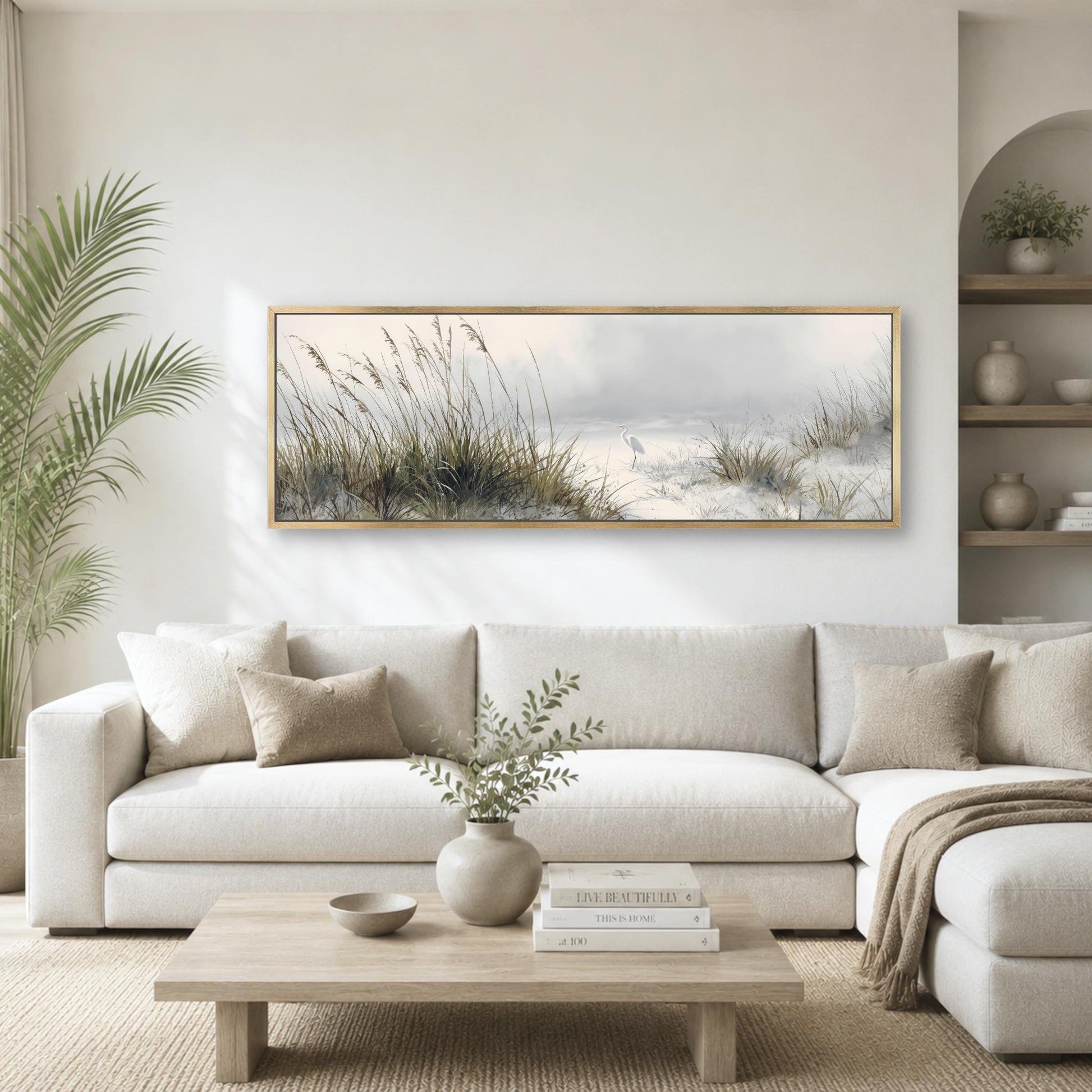 a serene living room with a large, framed painting of a beach scene hanging on the wall above a white sofa. The painting features tall grasses swaying in the breeze and a body of water in the background. The room is decorated with a wooden coffee table, a potted plant, and a vase with greenery, creating a cozy and inviting atmosphere.